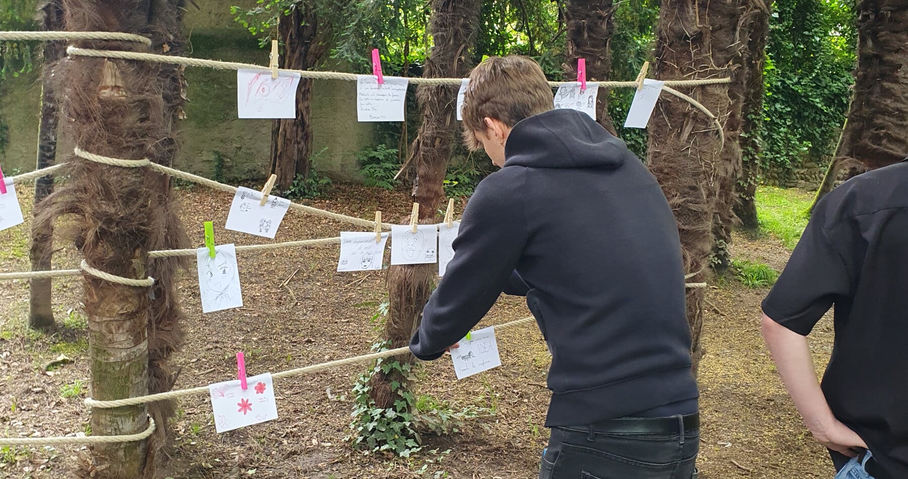 Un ragazzo, vestito completamente di nero, è fotografato di spalle mentre appende alcuni disegni a delle corde tese tra due alberi.