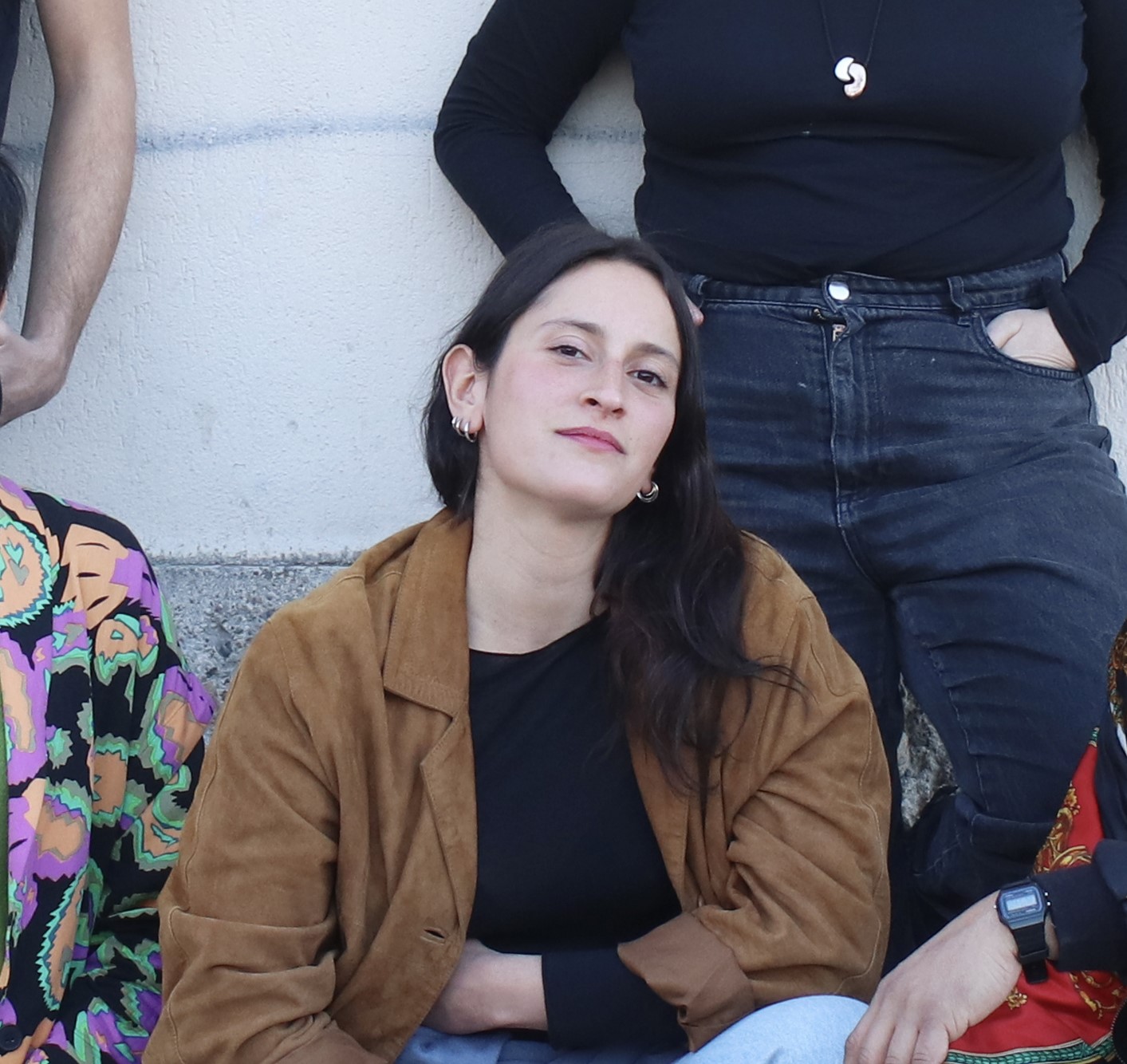 Alessandra seduta in mezzo a delle altre persone, indossa una giacca marrone e una maglia nera