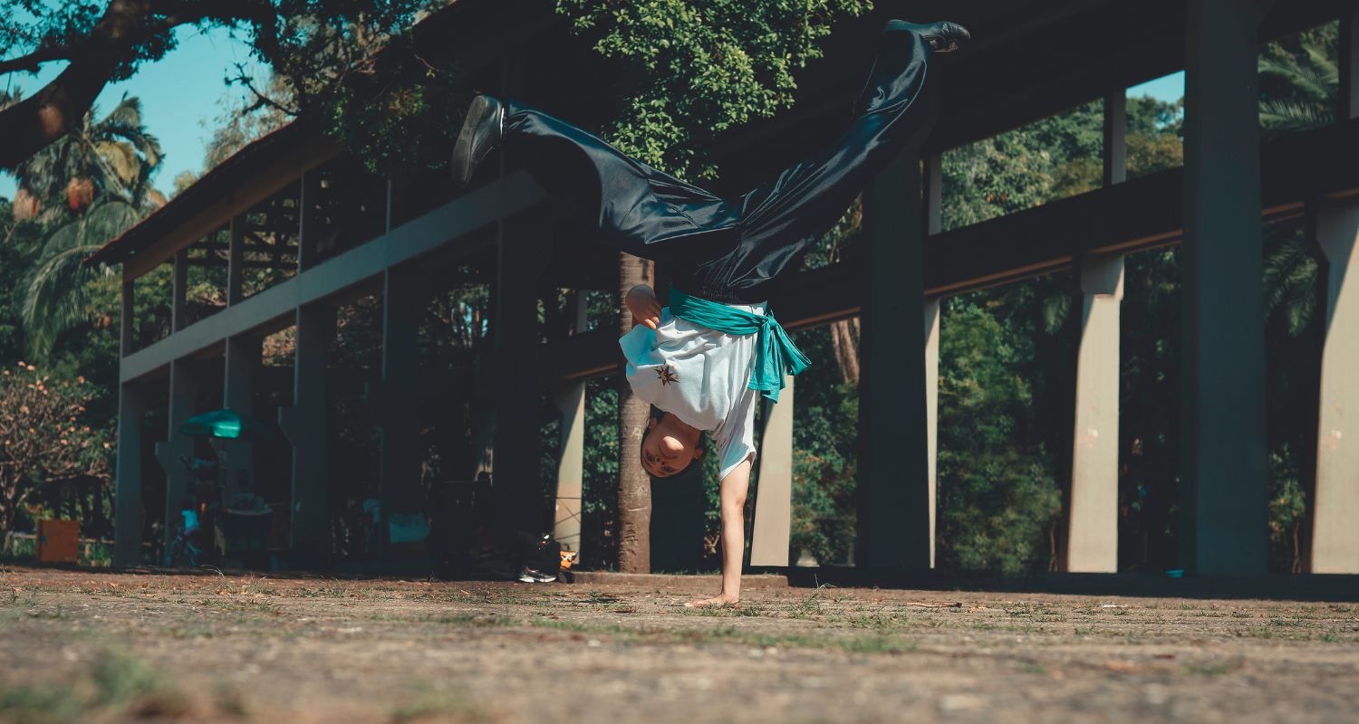 Una ragazza pratica una mossa di capoeira: è a testa in già e regge tutto il suo corpo con la sola mano sinistra. La gambe sono divaricate in aria.