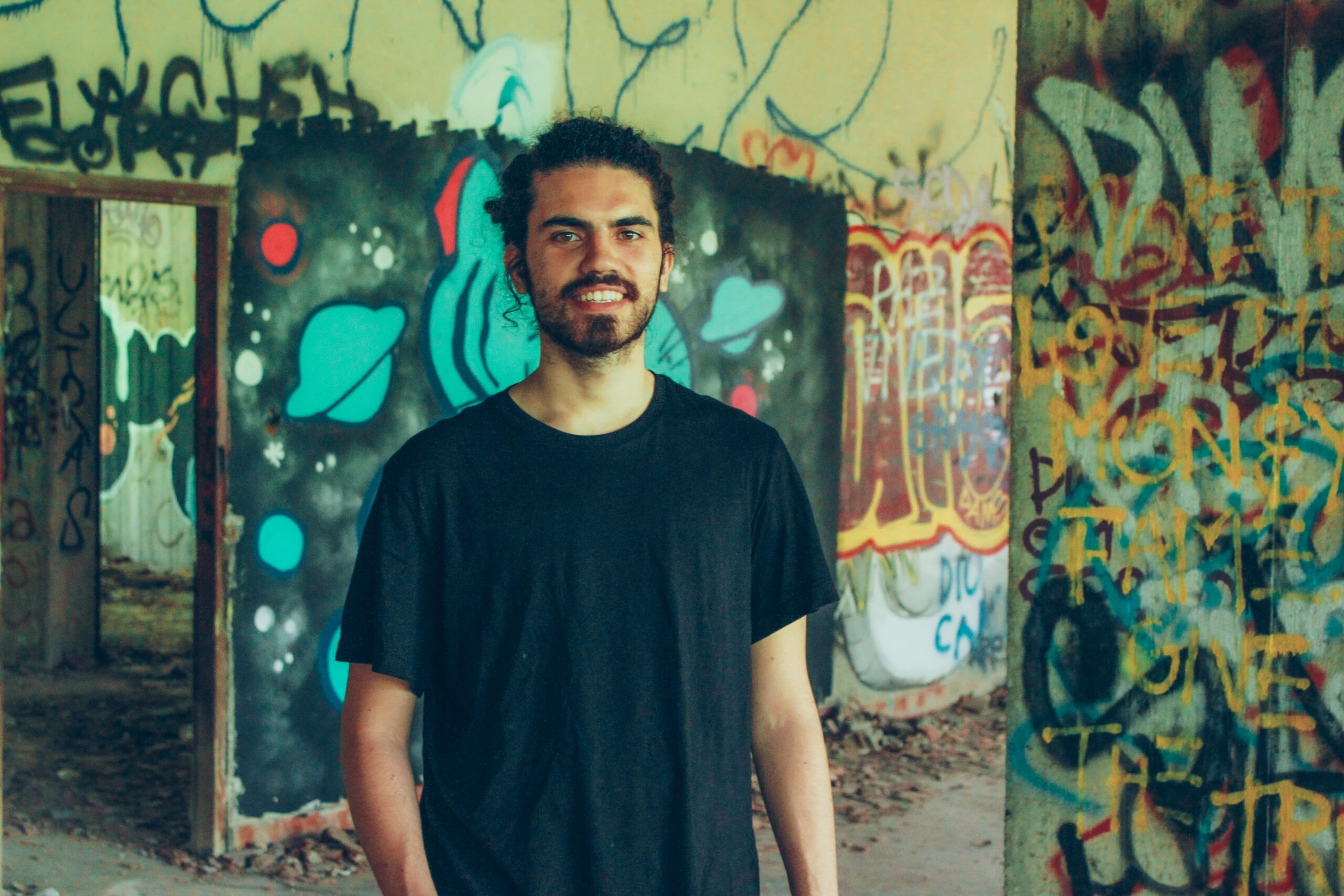 Giovanni è in piedi davanti a un edificio pieno di graffiti colorati. Indossa una maglietta nera e ha i capelli lunghi raccolti.