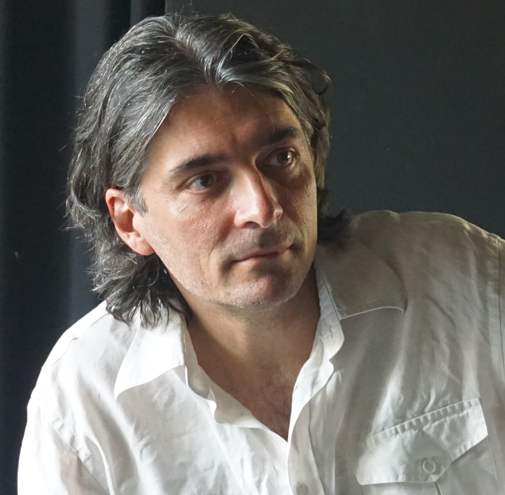 Damiano ripreso in primo piano, guarda fuori camera, verso destra. Ha una camicia bianca, lo sfondo è scuro, la luce arriva da sinistra.