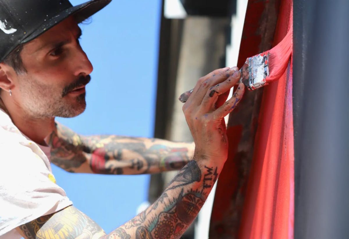Luca di profilo con una maglietta bianca e un cappellino nero, sta pitturando di rosso un muro con un pennello