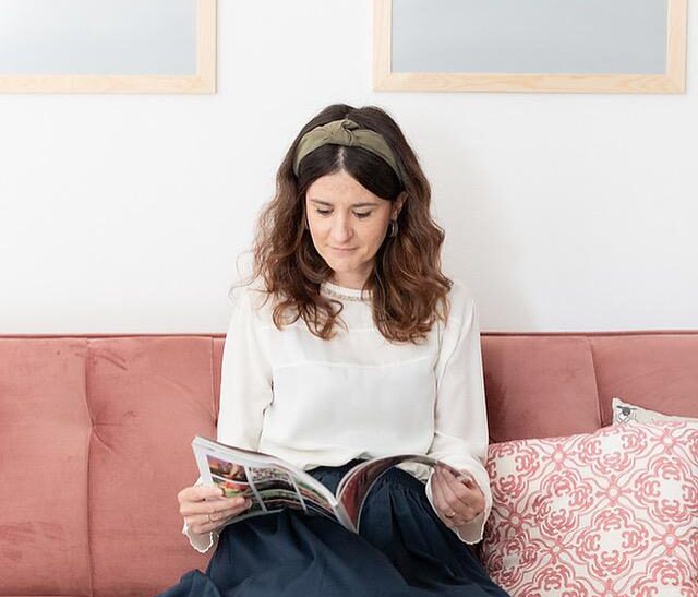 Nadia è seduta su un divano color mattone. Sfoglia un libro, indossa un cerchietto e una camicia chiara.