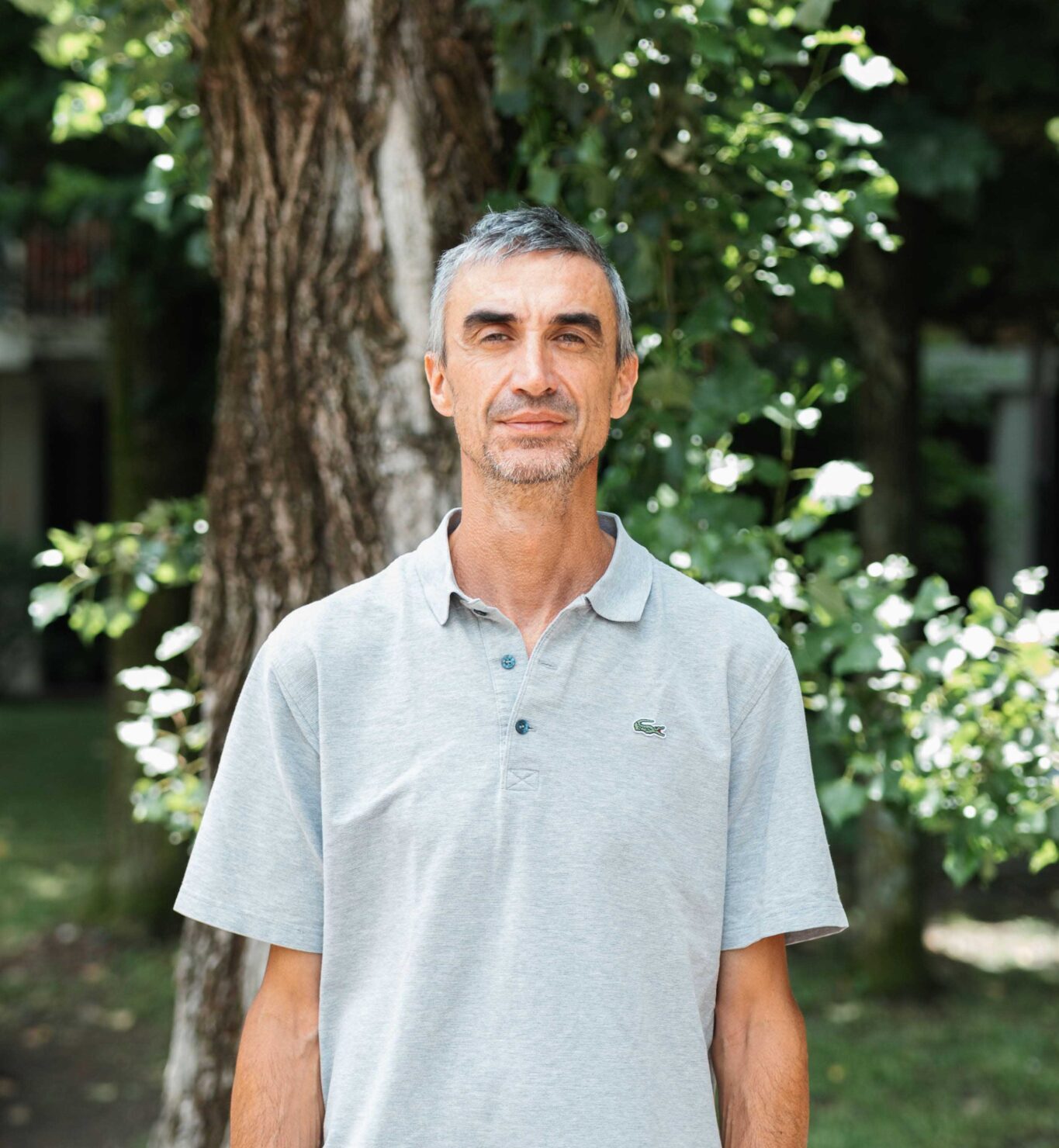 Paolo è in piedi davanti a un albero. Ha i capelli corti brizzolati e indossa una camicia a mani corte grigio chiaro.