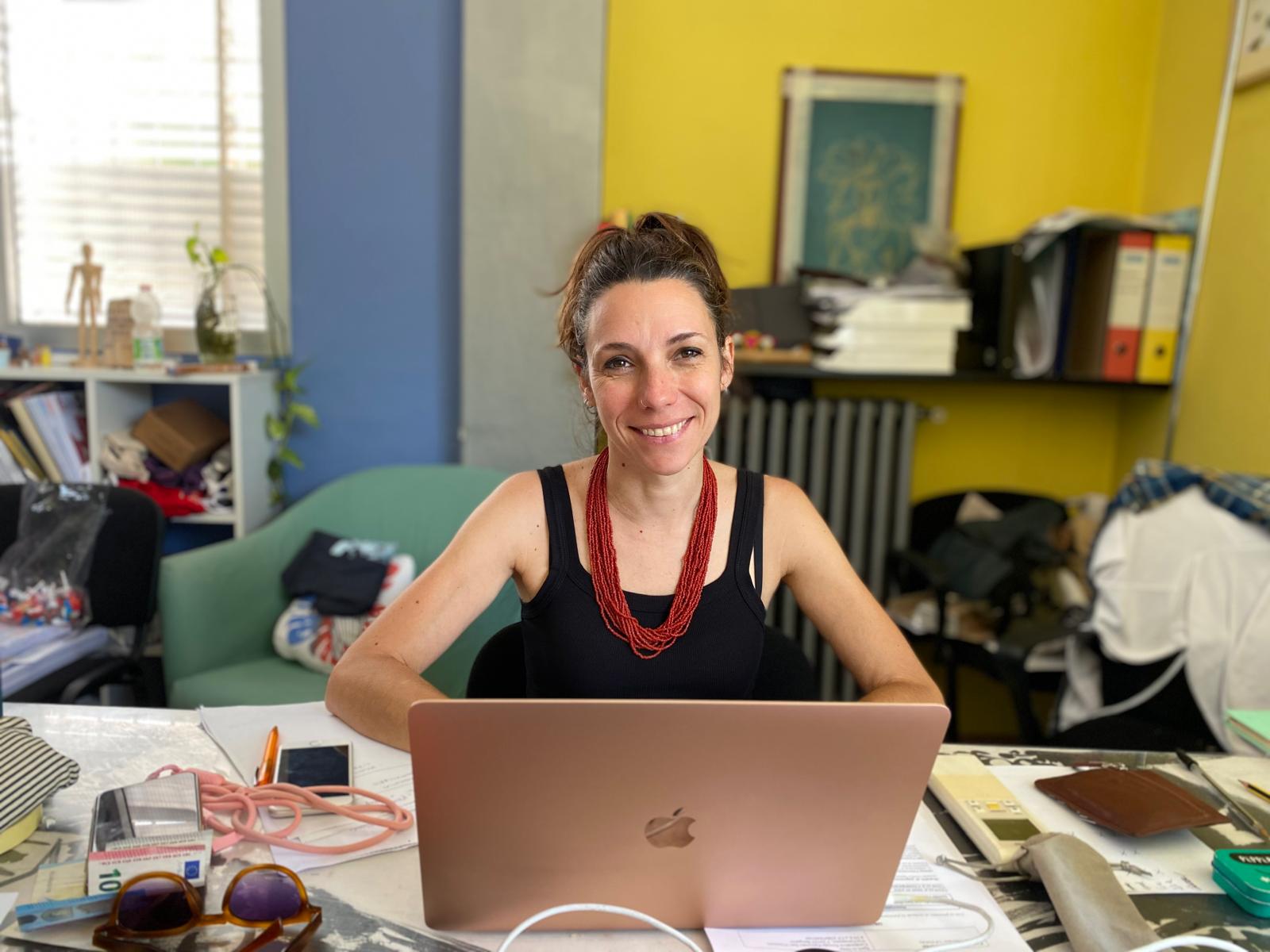 Simona è seduta davanti al computer portatile. Sorride e indossa una canottiera nera e una collana rossa