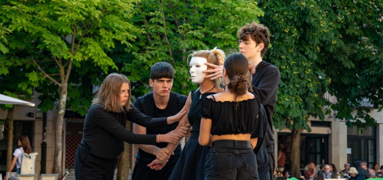Cinque persone vestite di nero in scena in uno spettacolo teatrale, all'aperto. La ragazza al centro indossa una maschera bianca.