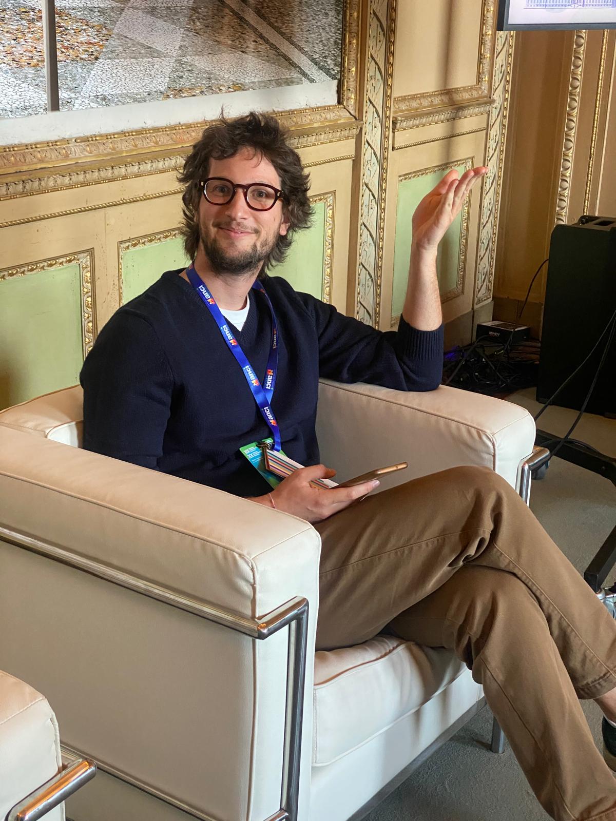 Nicolò seduto su una poltrona bianca, indossa un maglione blu. Porta gli occhiali, ha la barba e i capelli scuri.