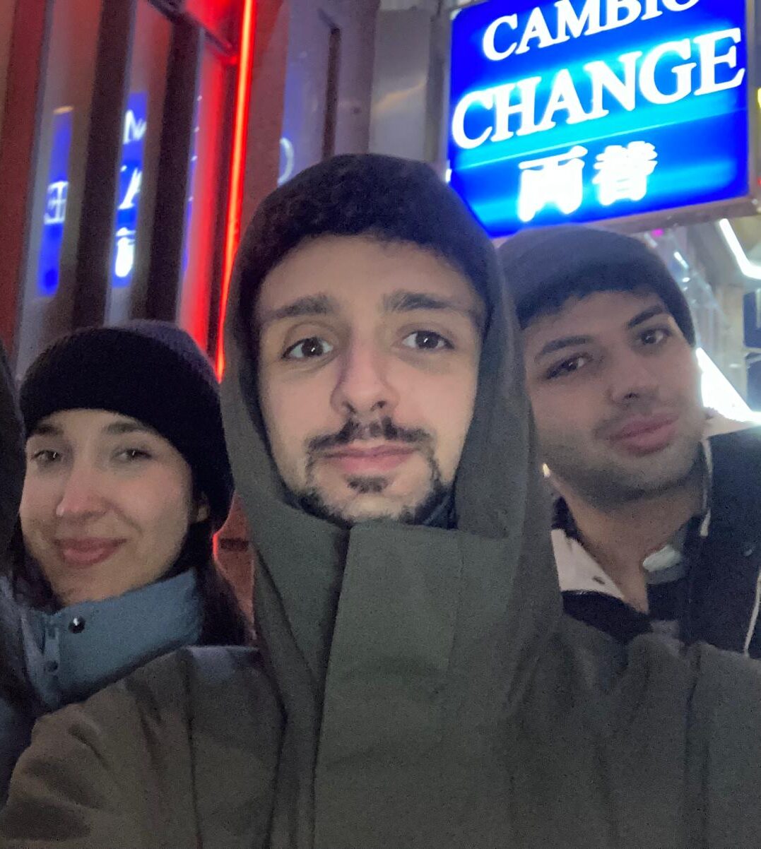 Matteo, al centro, fa un selfie con due amici, una ragazza sulla sinistra e un ragazzo a destra. Sullo sfondo un cartello al neon porta la scritta 