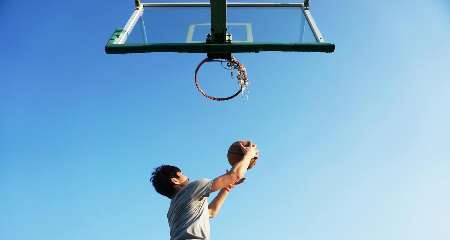 Un ragazzo sta saltando sotto canestro con un pallone da basket in mano. La foto è scattata dal basso.