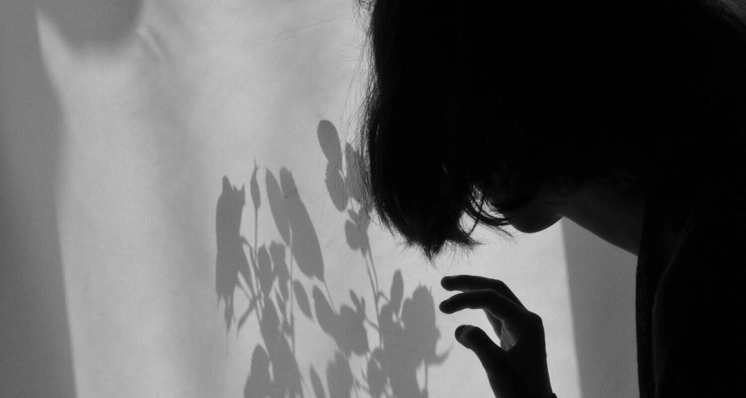 Foto in bianco e nero di una ragazza ripresa di spalle, girata per tre quarti, che tende la mano verso una parete su cui spicca l'ombra di una pianta in fiore. La foto è in bianco e nero ed è evocativa.