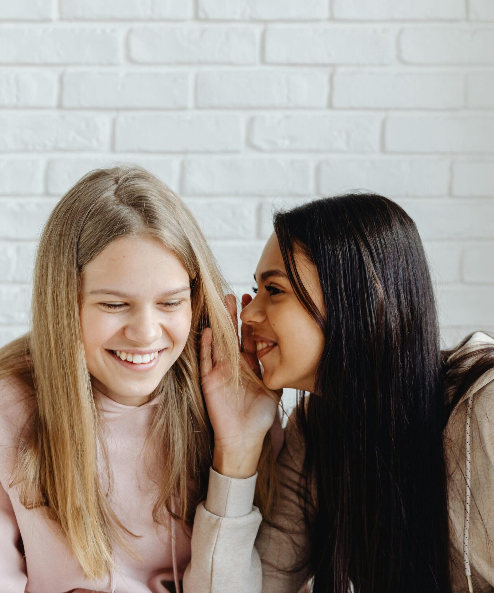 due teenager sorridono davanti a uno sfondo neutro. Una delle due parla all'orecchio dell'altra