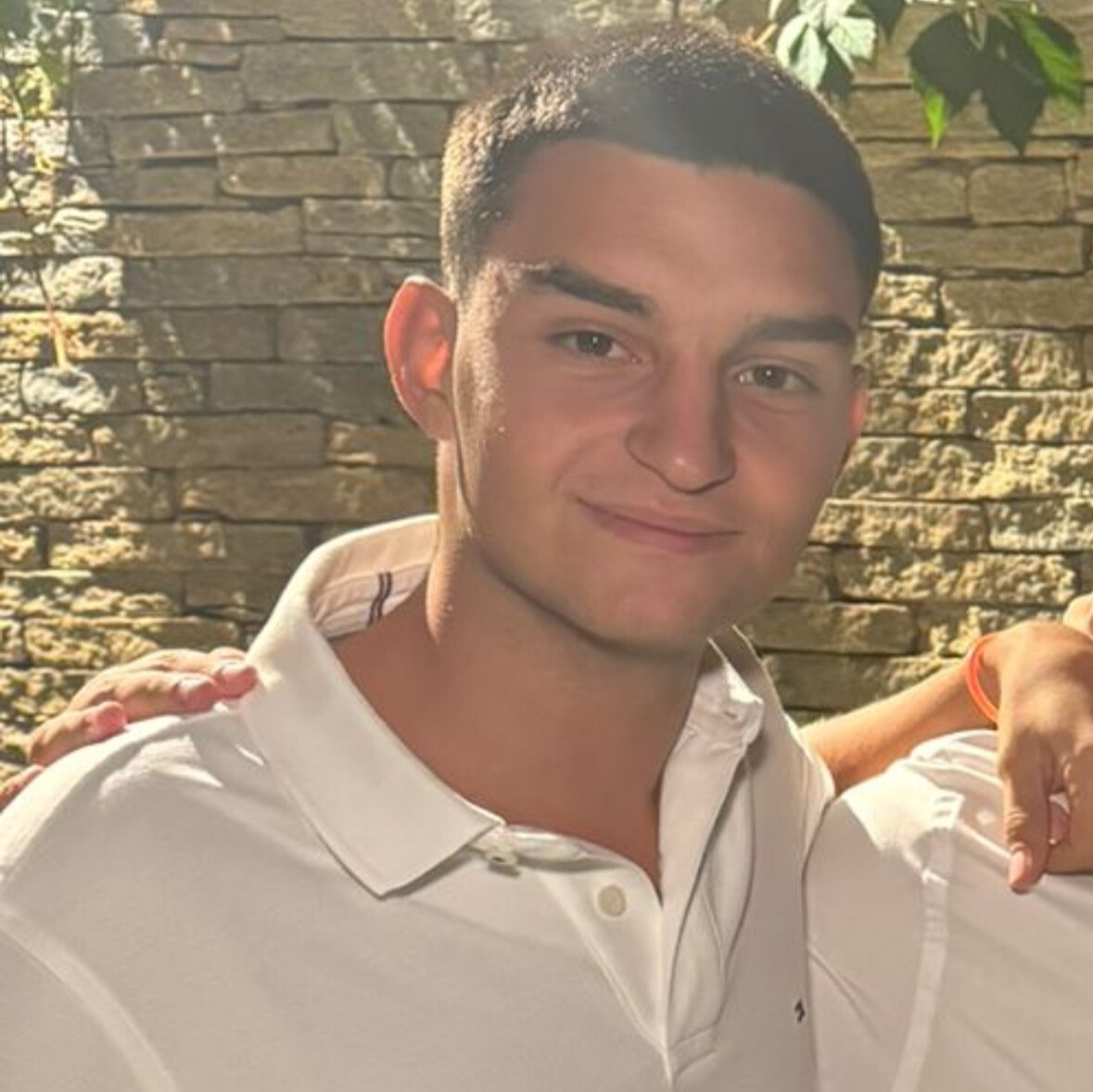 Andrea è in piedi, indossa una camicia a maniche corte bianca. Ha i capelli molto corti.
