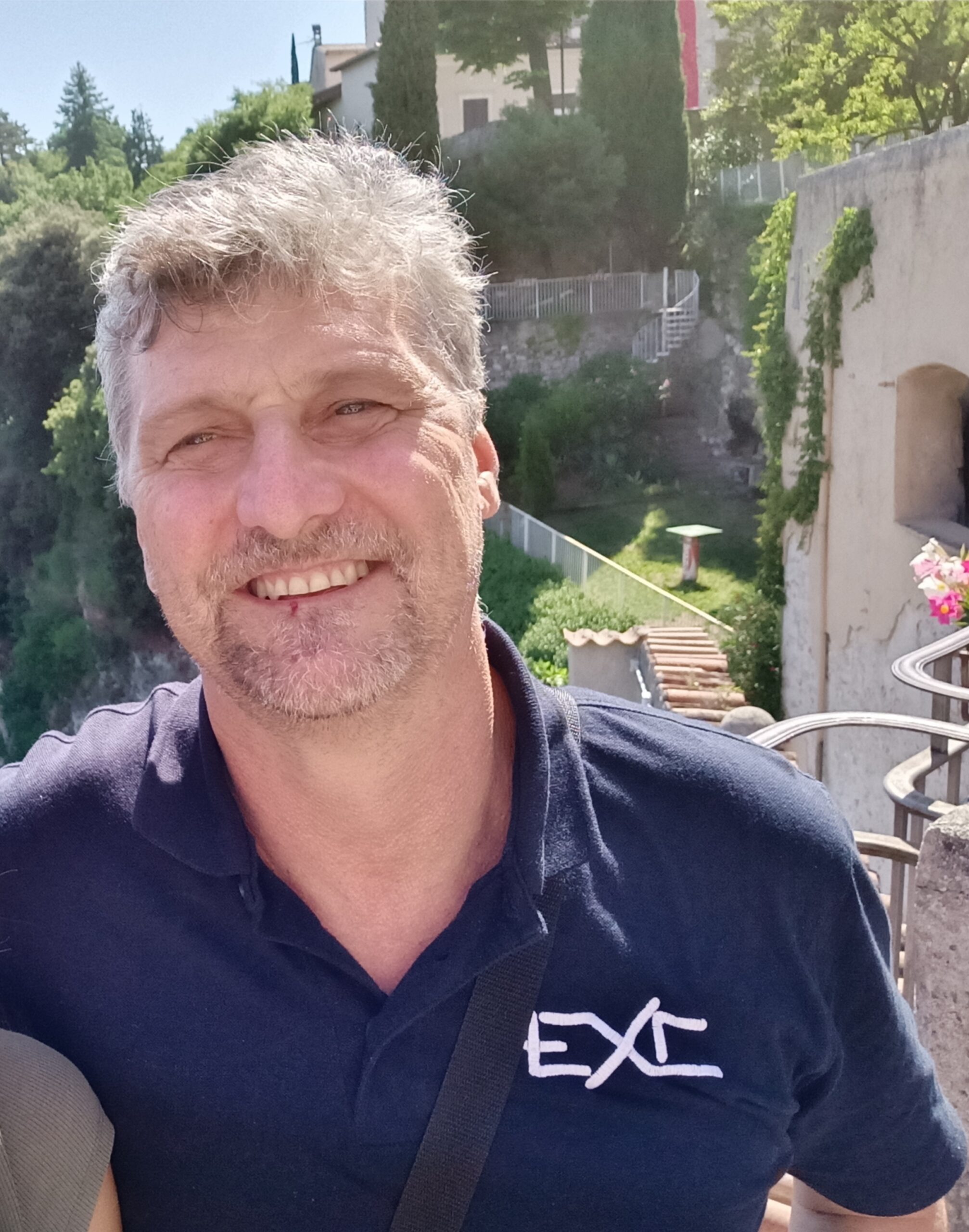 Primo piano di Alberto, sorride e ha una polo blu. Dietro di lui uno scorcio di casa e molti alberi