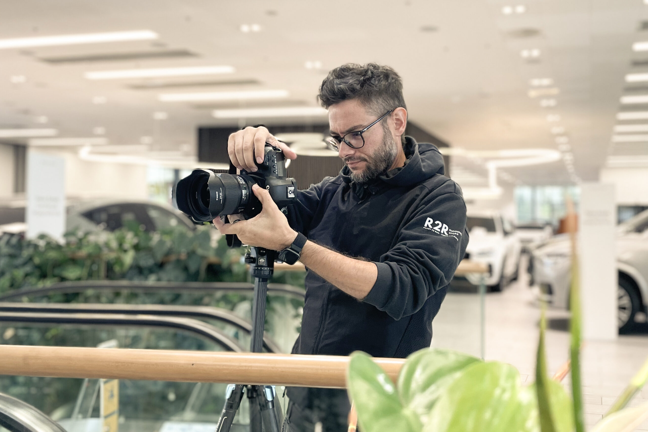 Stefano ha in mano la sua macchina fotografica e guarda dentro l'obiettivo che sta inquadrando, si trova all'interno di un luogo chiuso come un centro commerciale. Ha i capelli corti, gli occhiali e indossa una felpa nera.