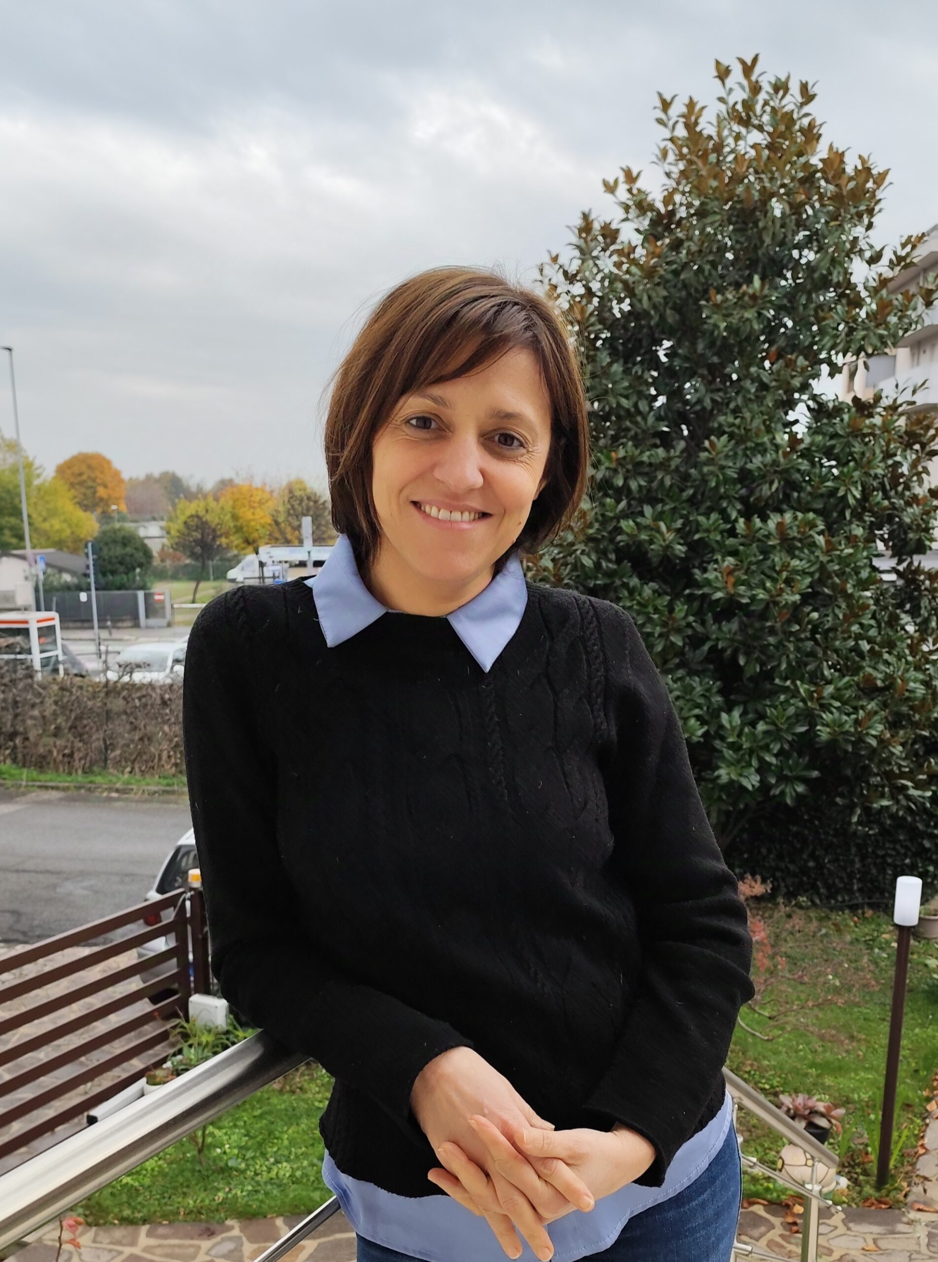 Anna di fronte a un panorama con un albero verde, ha un golfino scuro i capelli corti e scuri e sorride.