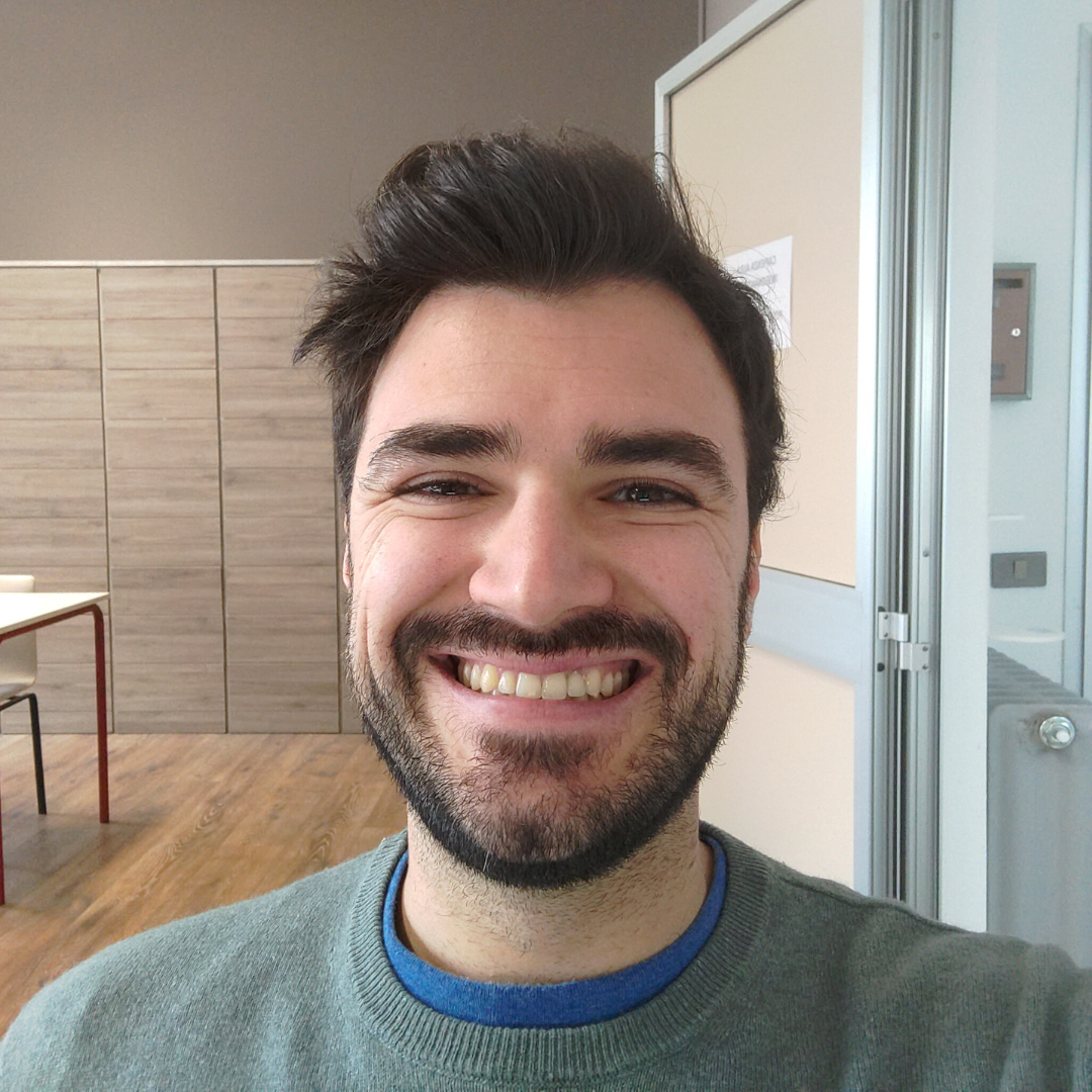 Marco sorride in un selfie. Ha i capelli scuri. Dietro di lui si vede un'aula di scuola