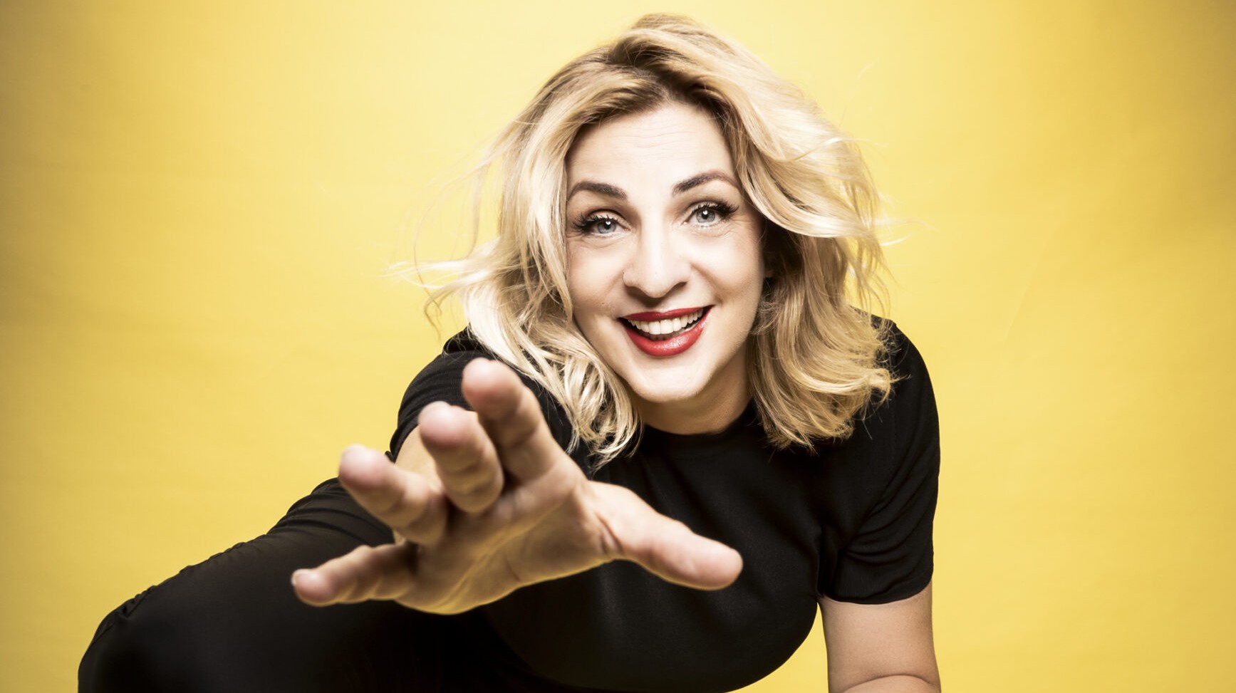 L'attrice è in posa: sorride in camera e tende la mano verso chi guarda. Lo sfondo è un set fotografico completamente giallo.