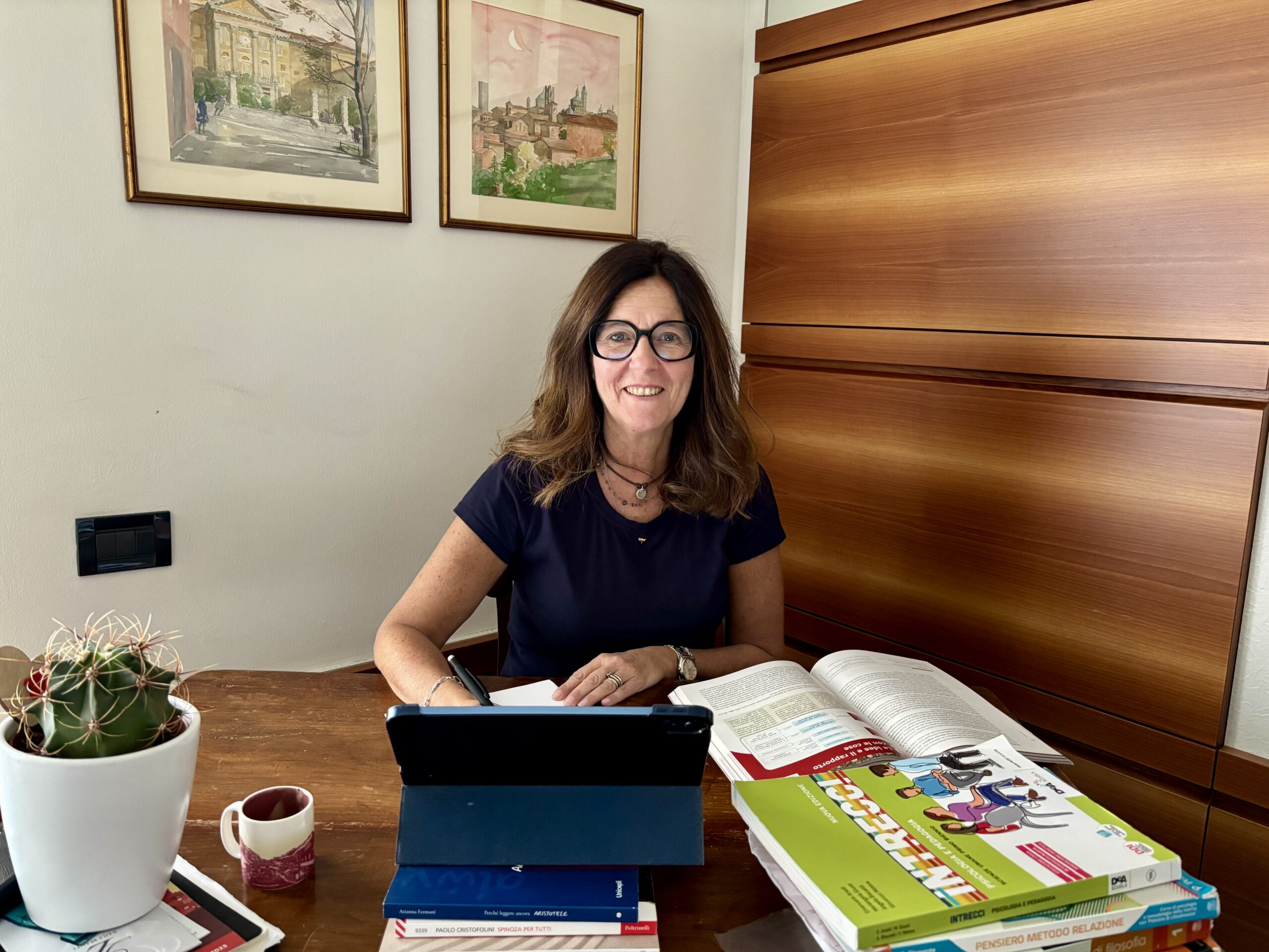 Francesca è seduta dietro a un tavolo dove ci osno dei libri, un tablet e una pianta e st ascrivendo su un foglio, indossa una maglia scura porta gli occhiali e ha i capelli lunghi e scuri.