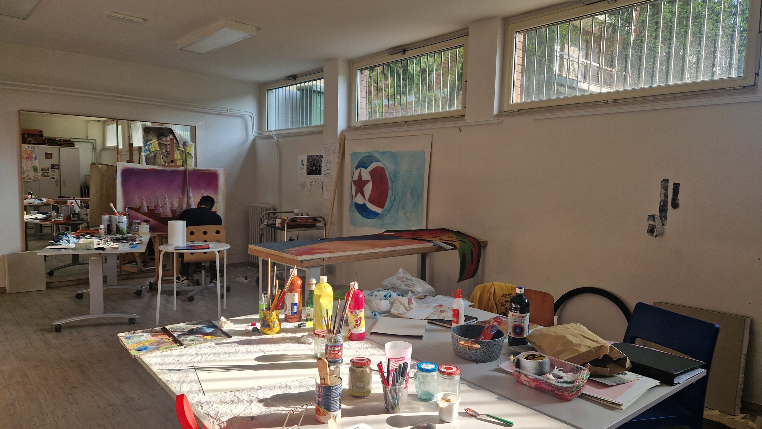 Foto di un artista all'opera in uno studio d'arte: in primo piano un tavolo con colori a tempera e pennelli, sullo sfondo l'artista che lavora a un grande quadro dalle linee geometriche.