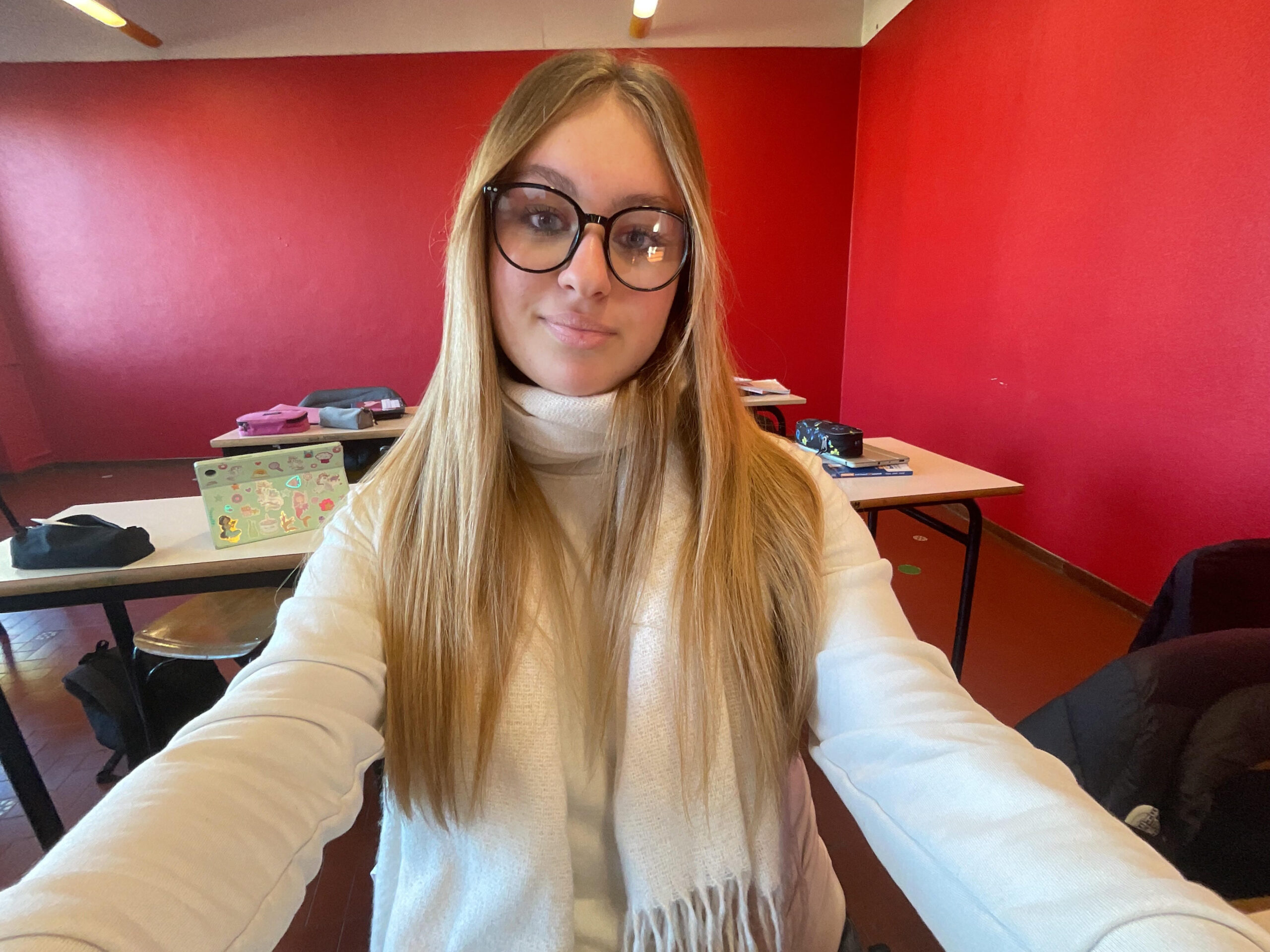 Sofia si sta facendo un selfie in un'aula con le pareti rosse. Ha i capelli lunghi e biondi, porta gli occhiali una sciarpa bianca e un maglione bianco.
