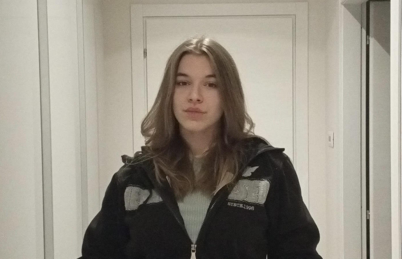 Karina è davanti a una porta bianca, indossa una felpa nera con la zip e scritte grigie aperta. Sotto indossa una maglietta grigia, ha i capelli lunghi e marroni.
