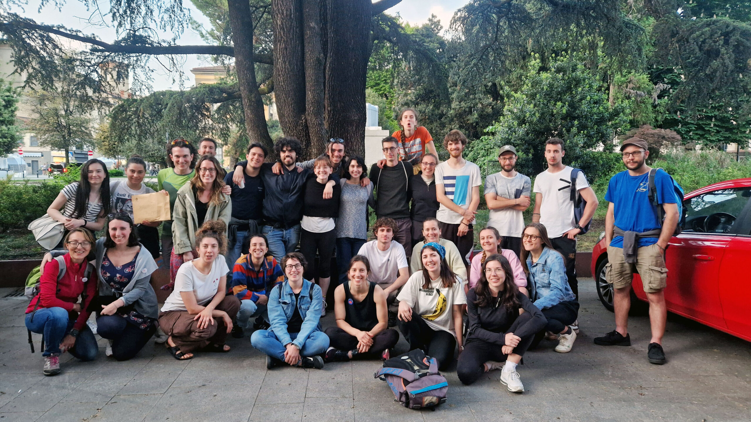 Gruppo numeroso di circa venticinque giovani posati insieme all’aperto, davanti a grandi alberi in un parco urbano. Alcuni sono in piedi e altri seduti o accovacciati a terra, con zaini e abbigliamento informale. L’atmosfera è sorridente e conviviale, come al termine di un’attività di gruppo o di un evento.
