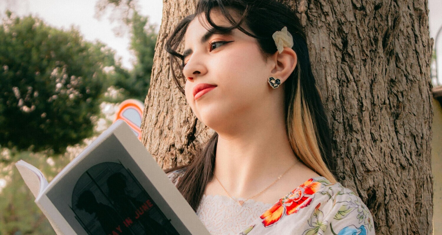 Una ragazza siede appoggiata al tronco di un albero, con un libro aperto in mano. Sembra persa nei propri pensieri.