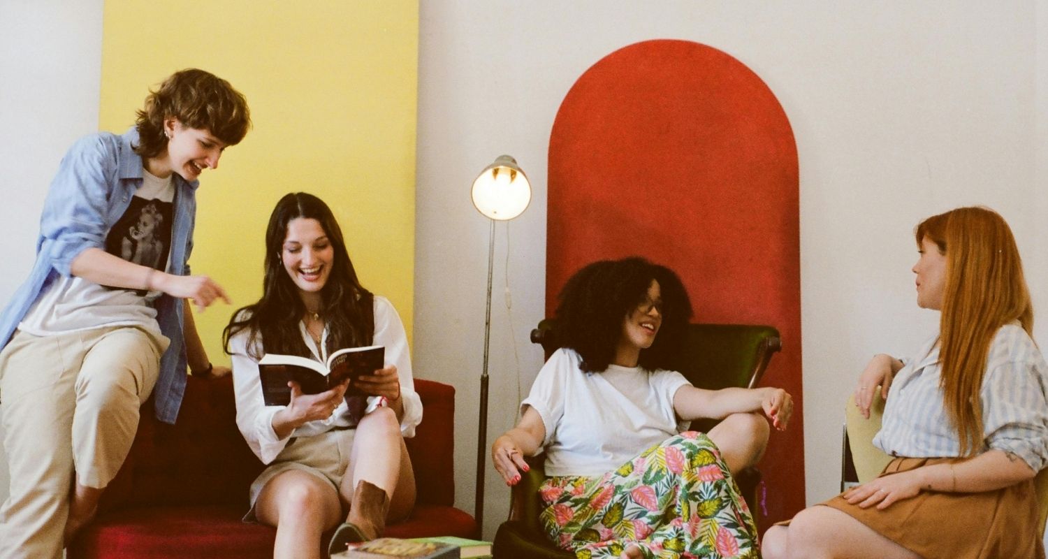 Quattro giovani sedute insieme in uno spazio colorato, mentre una ragazza legge ad alta voce da un libro e gli altri ascoltano e sorridono, creando un momento informale e conviviale.