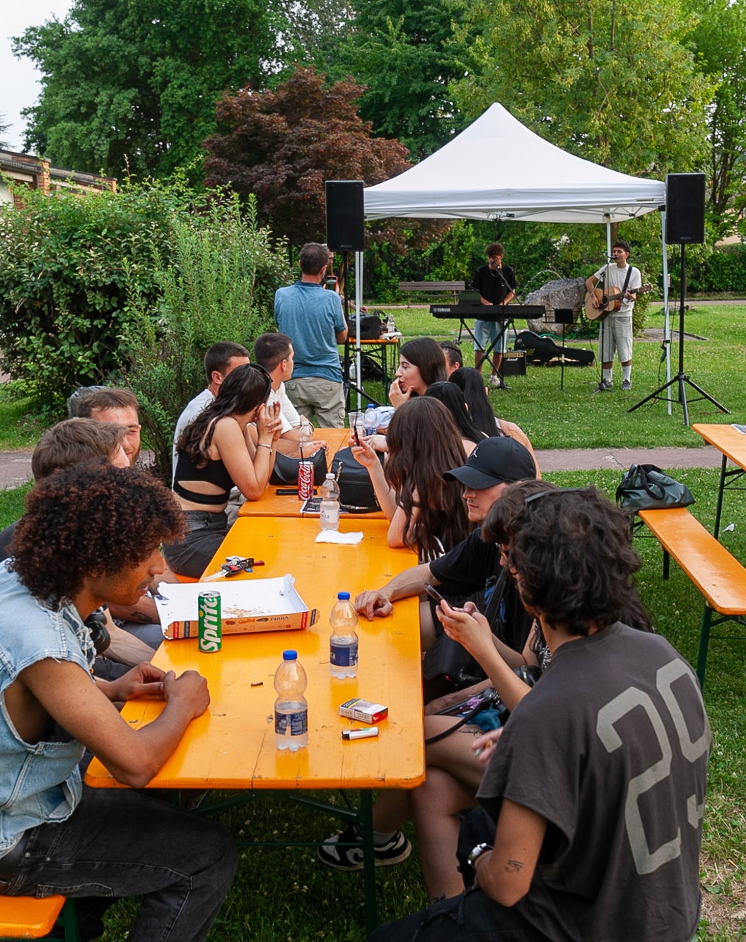 Gruppo di giovani seduti ai tavoli durante un evento all'aperto in un parco, mentre sullo sfondo due musicisti si esibiscono dal vivo sotto un gazebo creando un momento di incontro e convivialità.