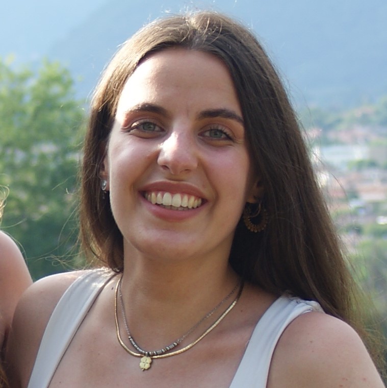 Chiara sorride, ha i capelli lunghi e castani e gli occhi chiari. Indossa una canottiera bianca e due collanine argento.