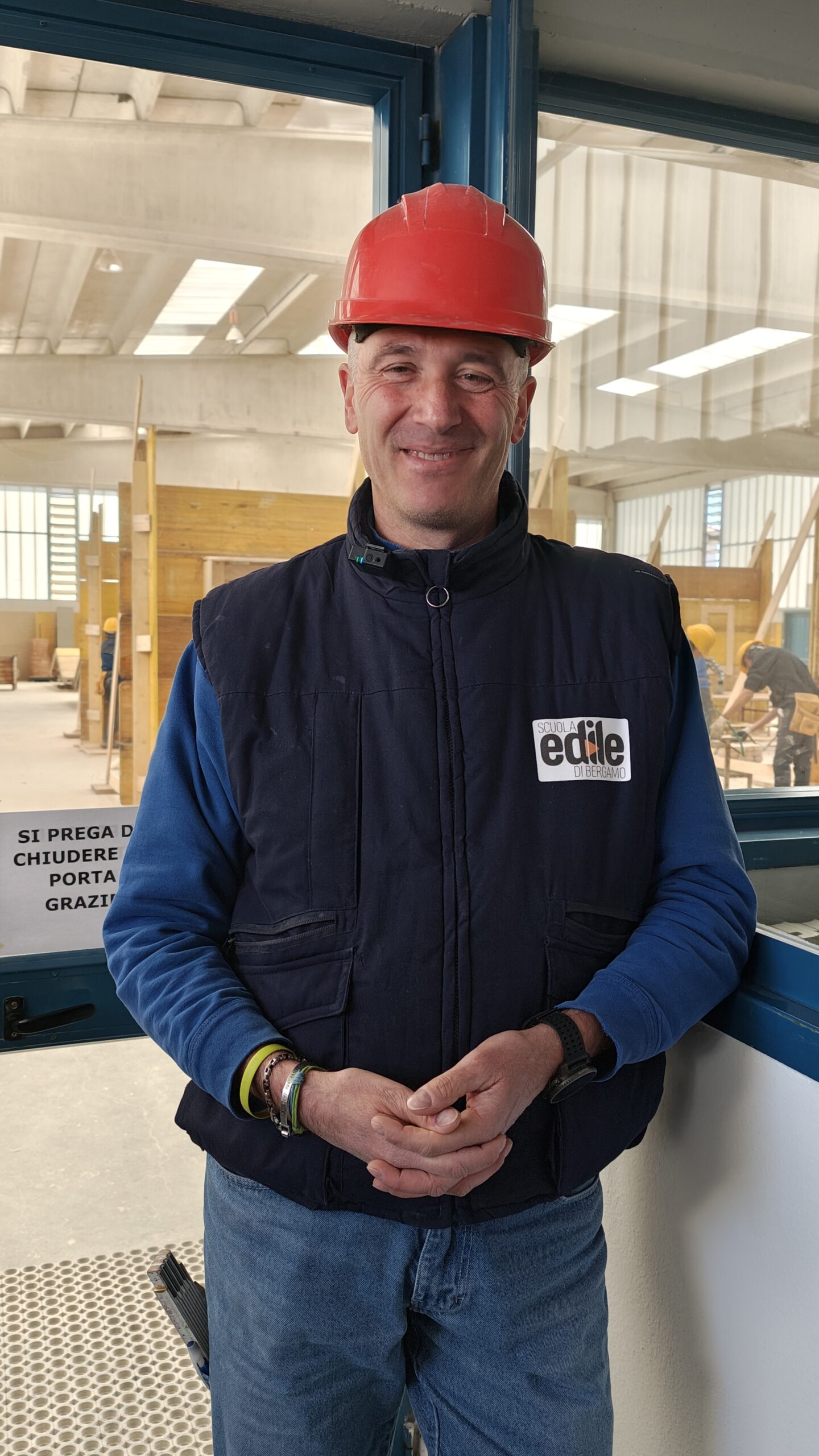Alessandro è in piedi, indossa un caschetto da cantiere rosso. Un gilet blu con il logo della Scuola edile, sotto una felpa blu e dei jeans.