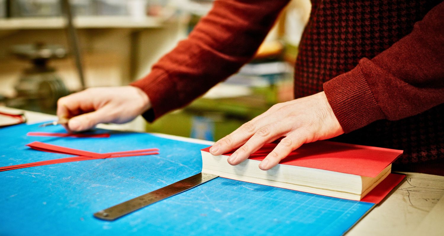 Persona che lavora alla rilegatura di un libro artigianale utilizzando carta rossa, taglierino e strumenti su un piano da lavoro.