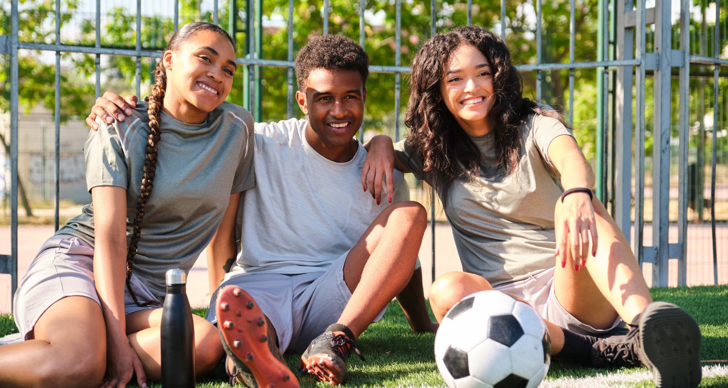 Tre ragazzi e ragazze seduti sull’erba di un campo da calcio, sorridenti e abbracciati, con un pallone e una borraccia davanti a loro, in un momento di pausa dopo aver giocato.