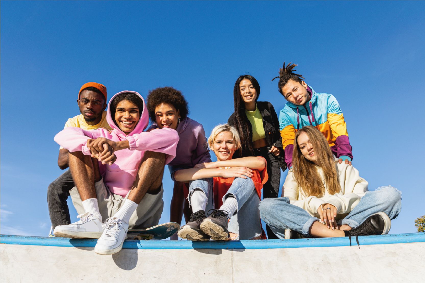 Gruppo di sei ragazzi e ragazze seduti insieme all’aperto su una struttura da skatepark, sorridenti e vestiti con abiti colorati casual, sotto un cielo azzurro limpido. L’immagine comunica inclusione, amicizia, creatività e partecipazione giovanile.