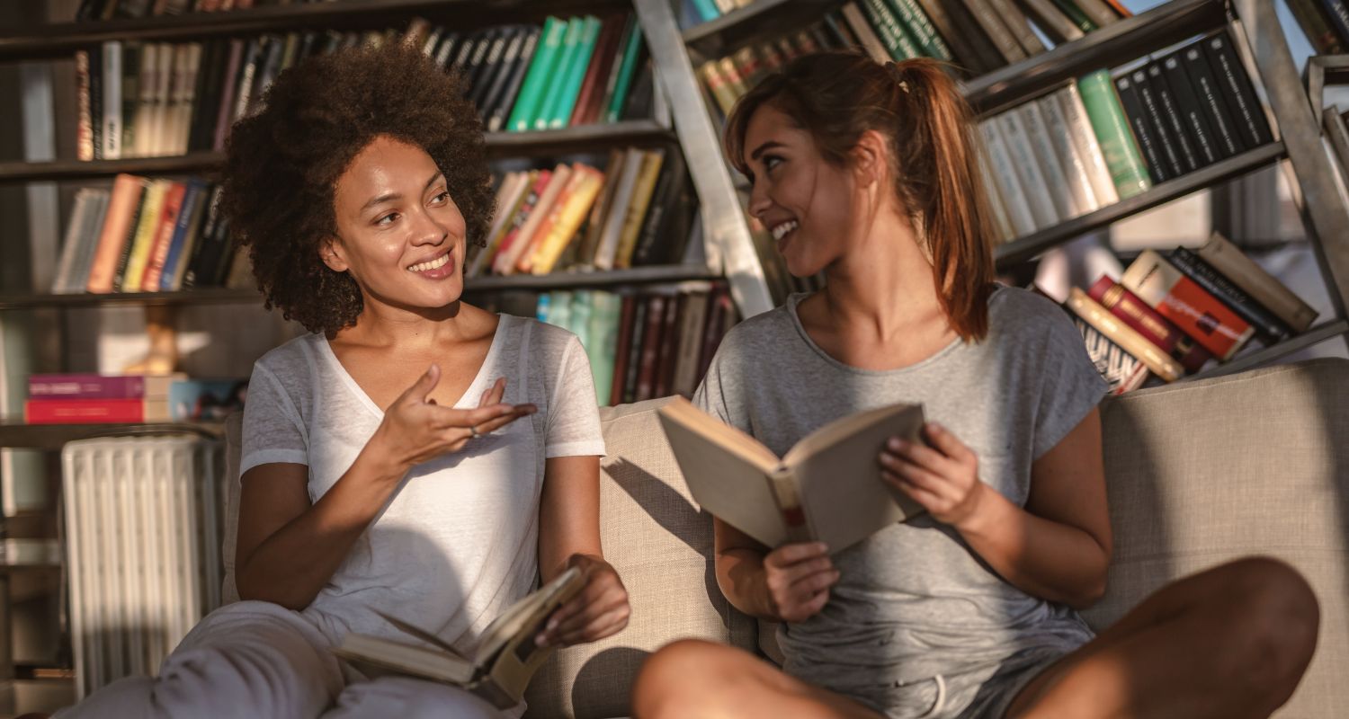Due ragazze sedute in una biblioteca chiacchierano sorridendo mentre leggono libri, circondate da scaffali pieni di volumi durante un incontro letterario informale.