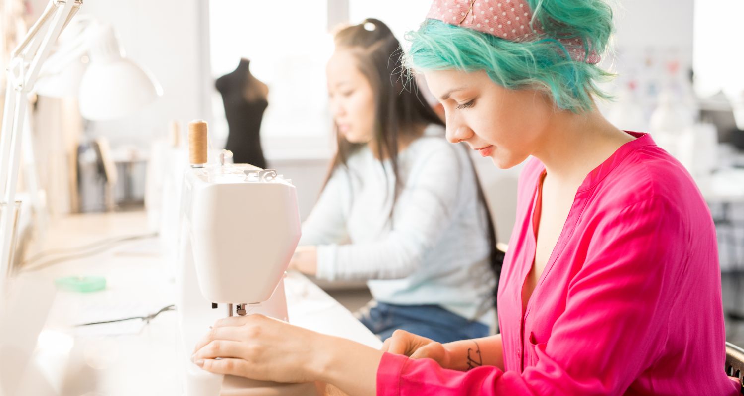 Due ragazze lavorano a un progetto di sartoria in un laboratorio luminoso, utilizzando una macchina da cucire e strumenti per cucito e personalizzazione di abiti.