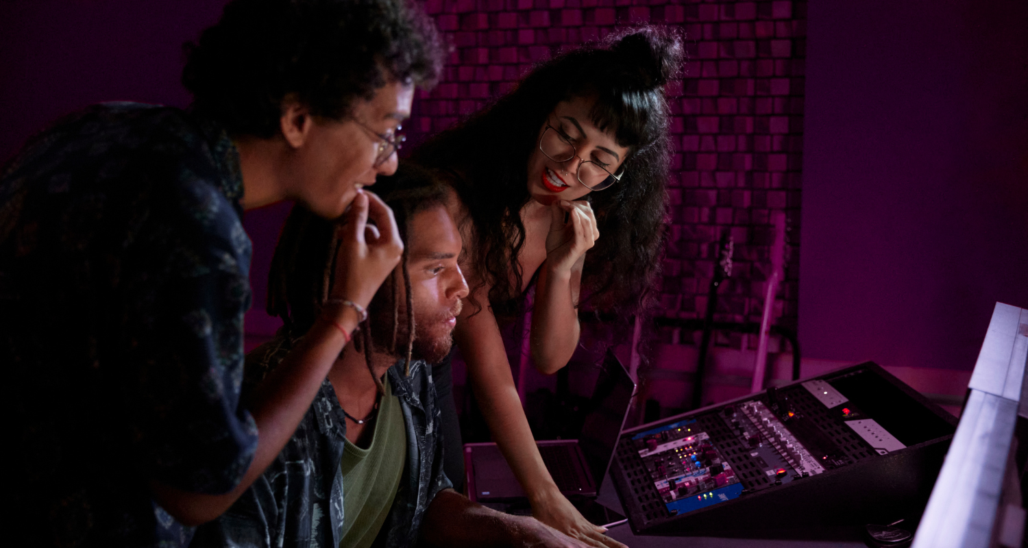 Tre giovani in uno studio musicale osservano insieme uno schermo e una console audio illuminata da luci viola e rosa, mentre lavorano a una produzione musicale.