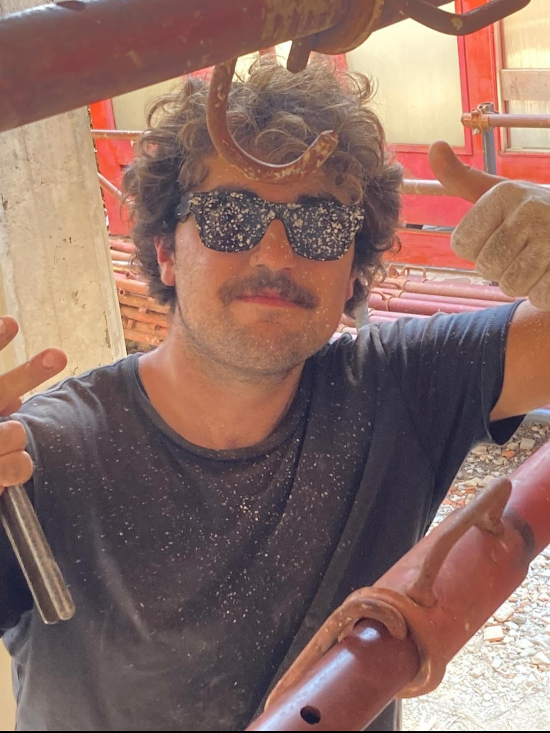 Davide è in piedi appoggiato ad un tubo. Indossa occhiali da sole scuri e sporchi di pittura bianca. Ha i capelli corti e castani. Indossa una maglietta a maniche corte blu e sporca di polvere. Si trova in un cantiere mentre lavora.