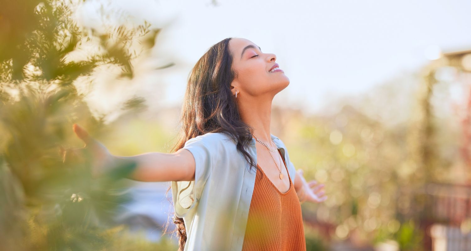 Donna all'aperto con gli occhi chiusi e le braccia aperte, immersa nella luce del sole. L'espressione rilassata e il contesto naturale trasmettono una sensazione di benessere, calma e connessione con l'ambiente circostante.