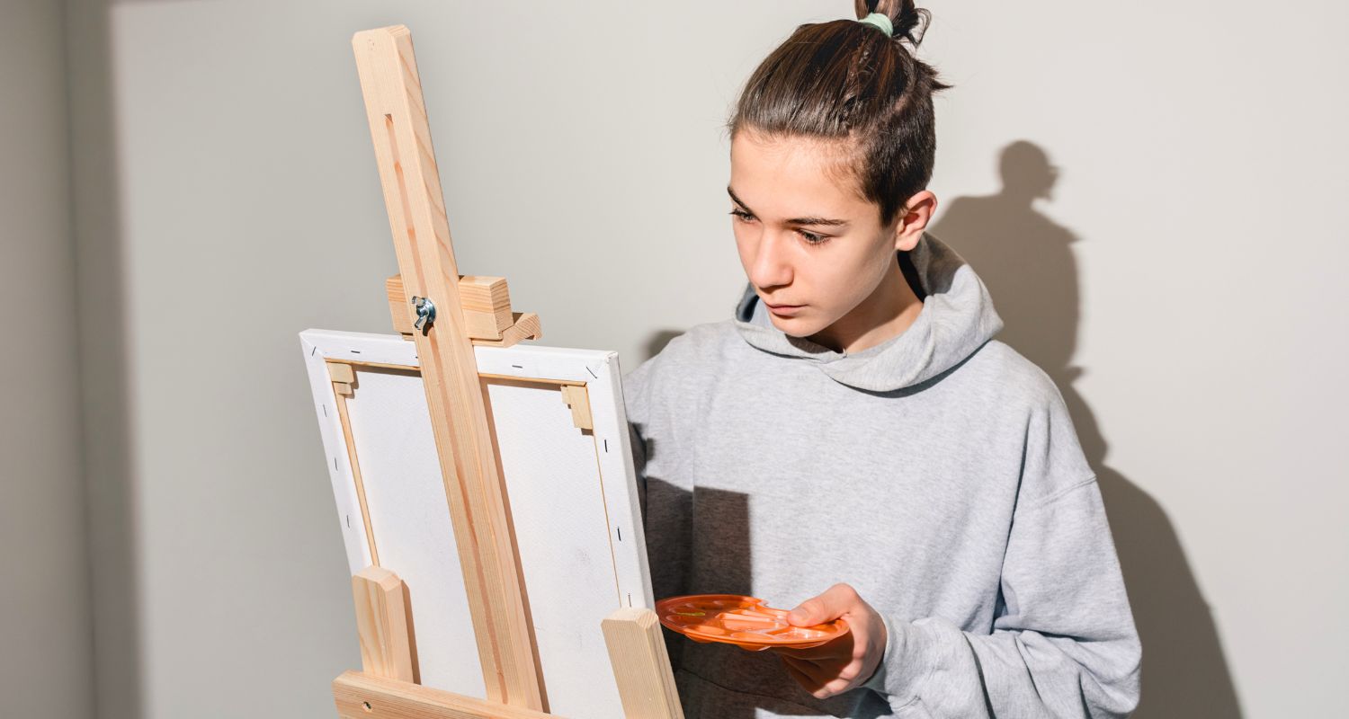 Giovane impegnato in un’attività artistica davanti a un cavalletto, mentre osserva attentamente una tela e tiene in mano una tavolozza con i colori. La scena richiama creatività, concentrazione e libera espressione attraverso l’arte.