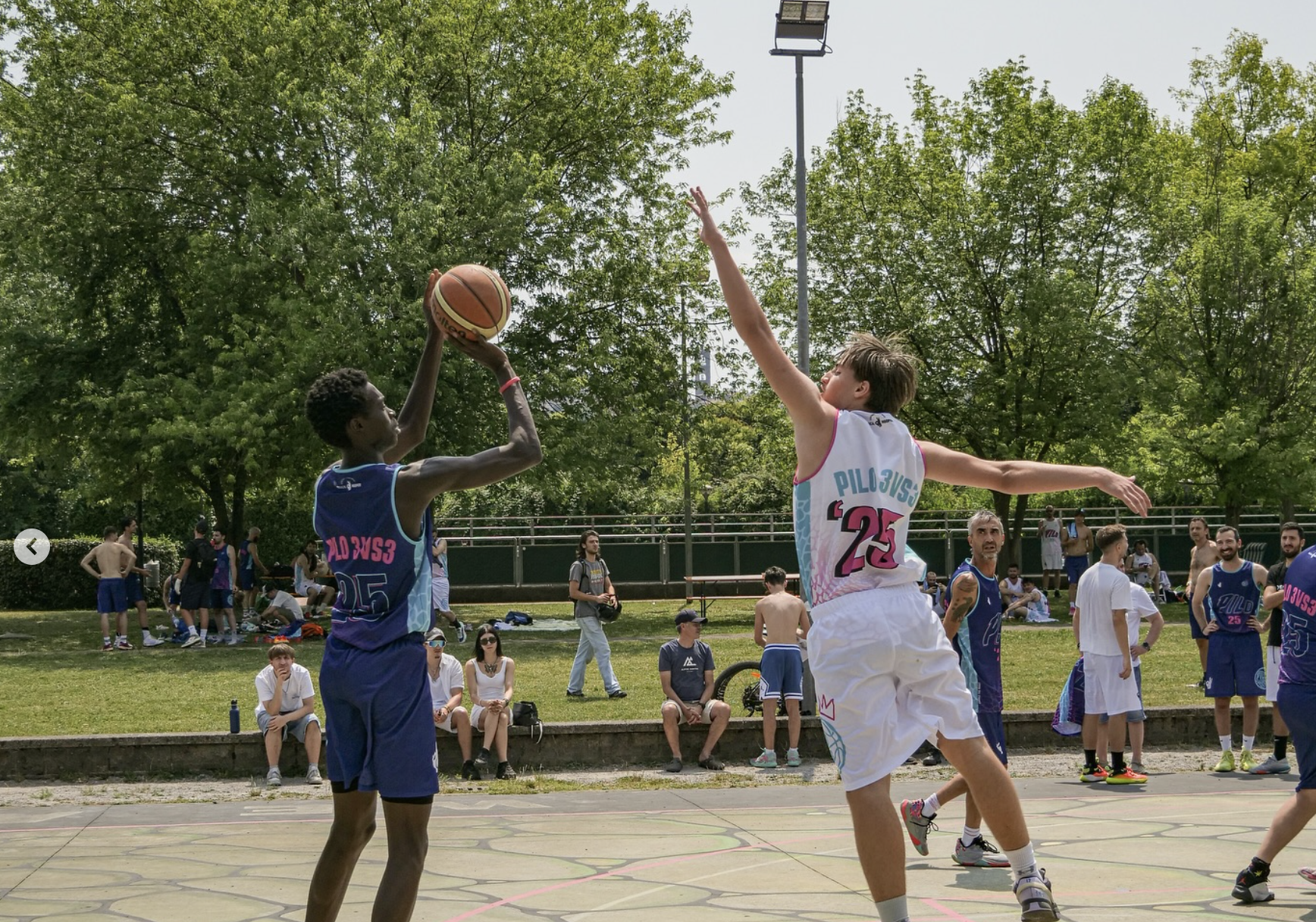 Partita di basket 3 contro 3 all'aperto durante il Torneo del Pilo. Un giocatore in maglia blu sta effettuando un tiro mentre un avversario in maglia bianca tenta di contrastarlo. Sullo sfondo spettatori e altre squadre seguono le partite nel campo del Centro Sportivo R. Pilo di Bergamo.