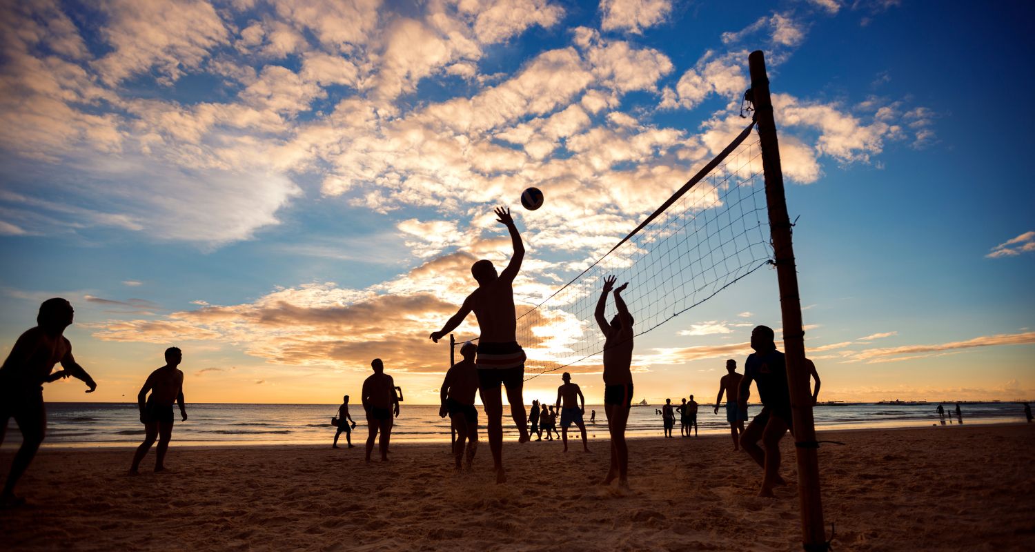 Un gruppo di ragazze e ragazzi gioca a pallavolo su un campo all'aperto al tramonto. Una persona salta per schiacciare la palla mentre altre sono pronte a riceverla, con il mare e il cielo illuminato dai colori del tramonto sullo sfondo.