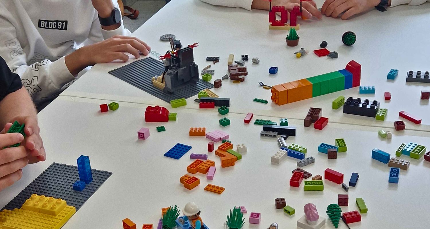 Giovani partecipanti seduti attorno a un tavolo mentre costruiscono con mattoncini LEGO colorati durante un'attività laboratoriale collaborativa. Sul tavolo sono presenti diverse costruzioni, basi di montaggio e numerosi pezzi sparsi utilizzati per un esercizio creativo di gruppo.