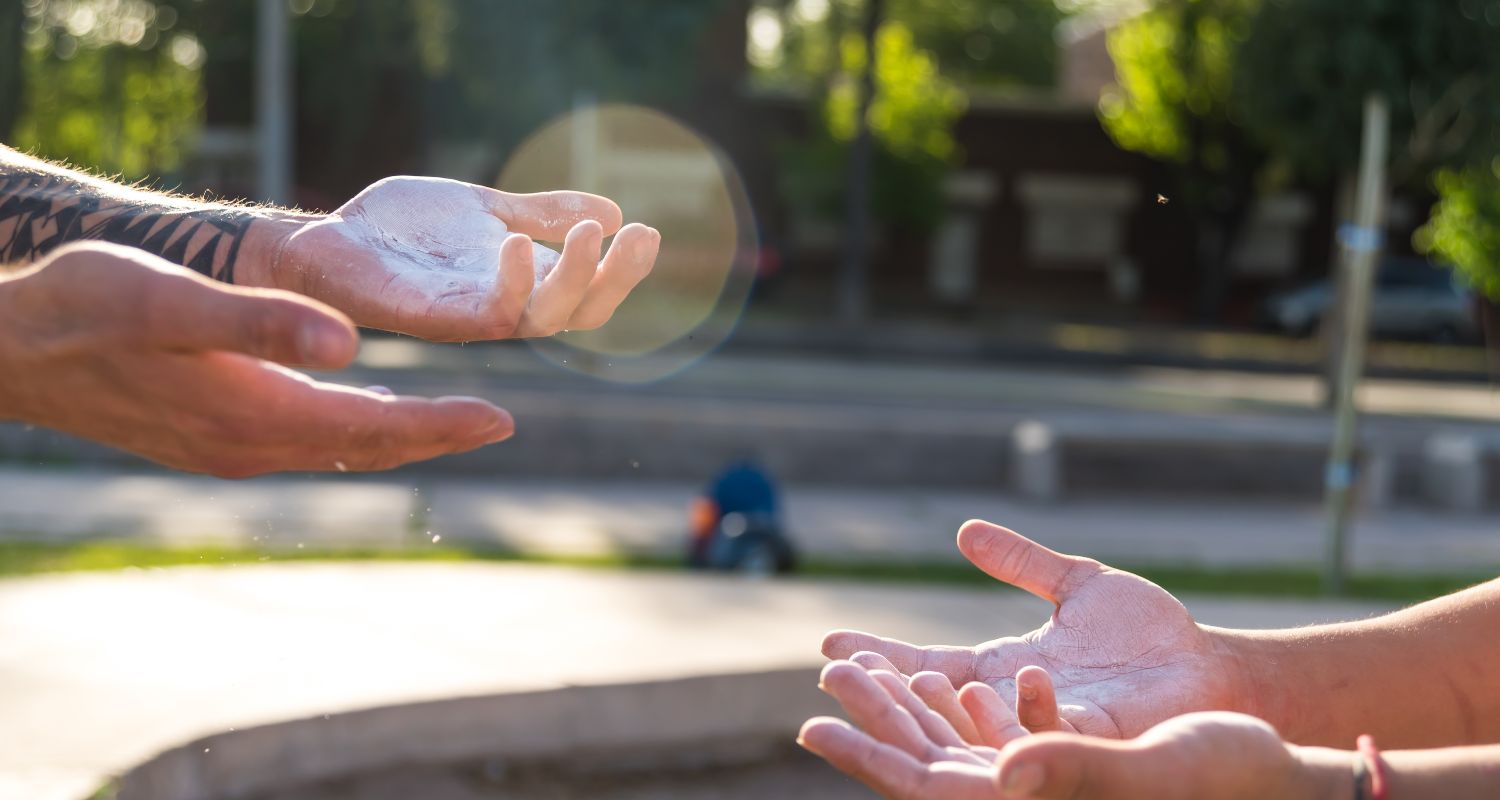 Primo piano di due persone che si preparano a praticare street workout all'aperto, con le mani ricoperte di magnesite. Sullo sfondo sfocato si intravede l'area di allenamento del Parco Goisis, illuminata dalla luce del tardo pomeriggio