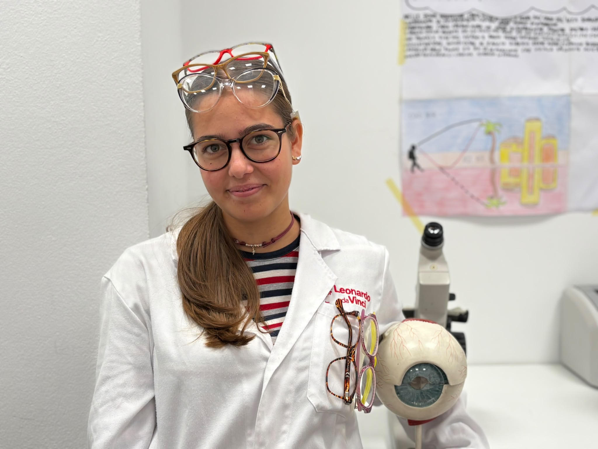 Marianna porta degli occhiali da vista neri. Ha i capelli lunghi, castano chiaro e indossa un camice bianco. Accanto a lei c'è un modellino di un occhio con accanto altri occhiali. Sopra la fronte indossa anche altri occhiali da vista di diversi colori.