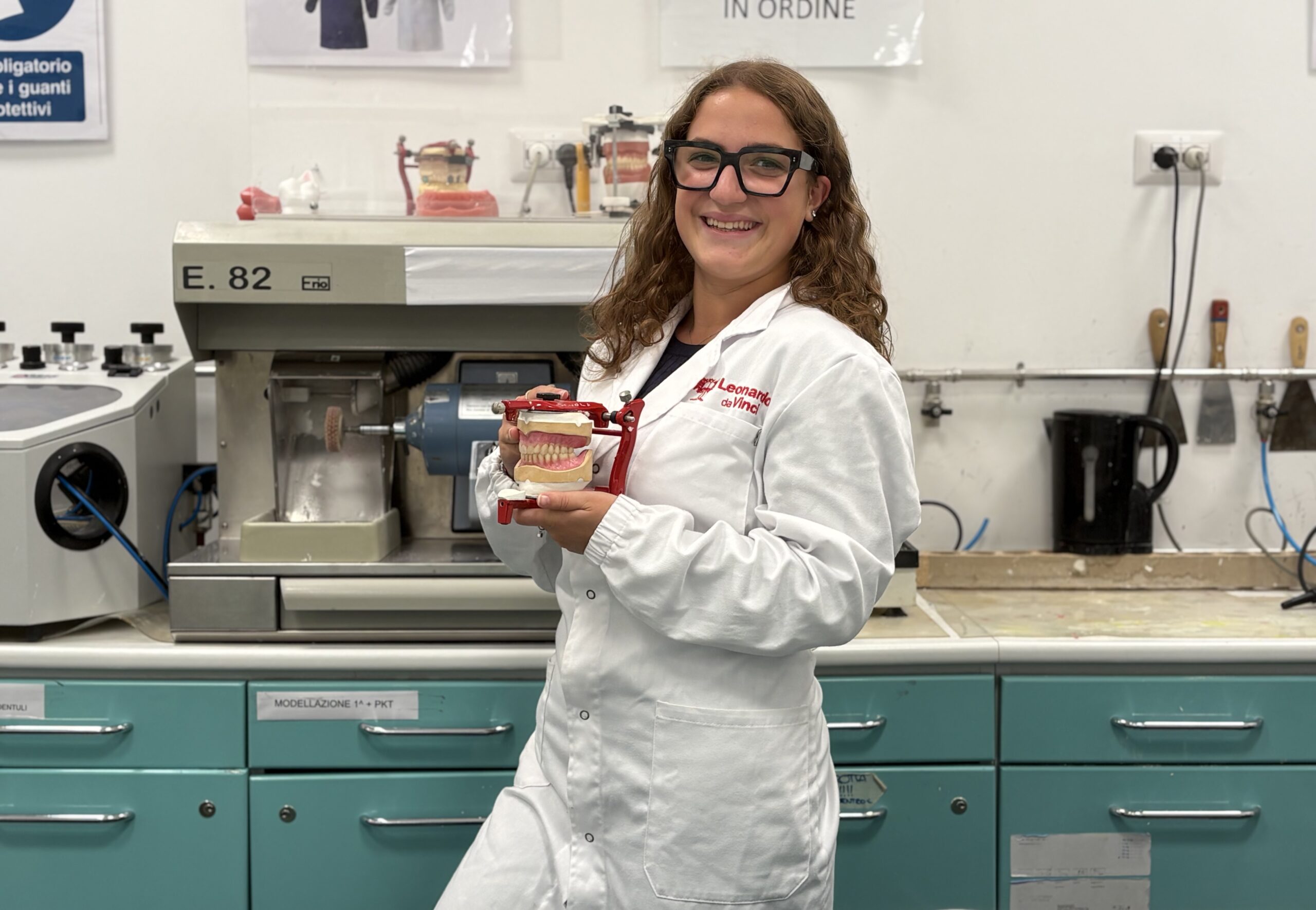 Francesca indossa un camice lungo bianco, e porta degli occhiali da vista neri. Ha i capelli lunghi e biondi e sorride. In mano a un modellino di una bocca con i denti. Sulla sfondo si trovano diversi oggetti del laboratorio in cui si trova