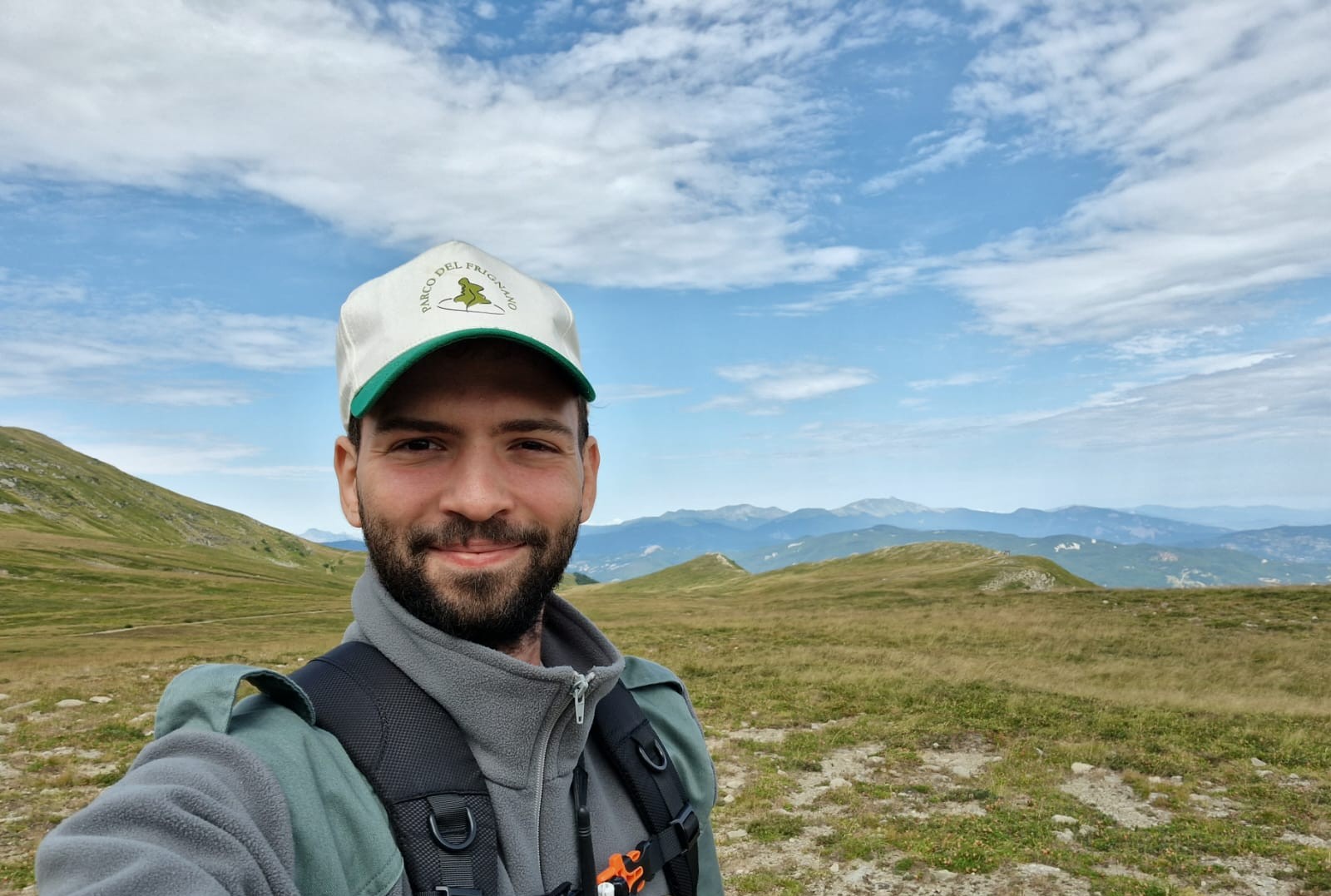 Filippo, indossa un cappellino bianco e un pile grigio e verde. Ha la barba corta e scura. Dietro di lui c'è il panorama di un prato con delle montagne.