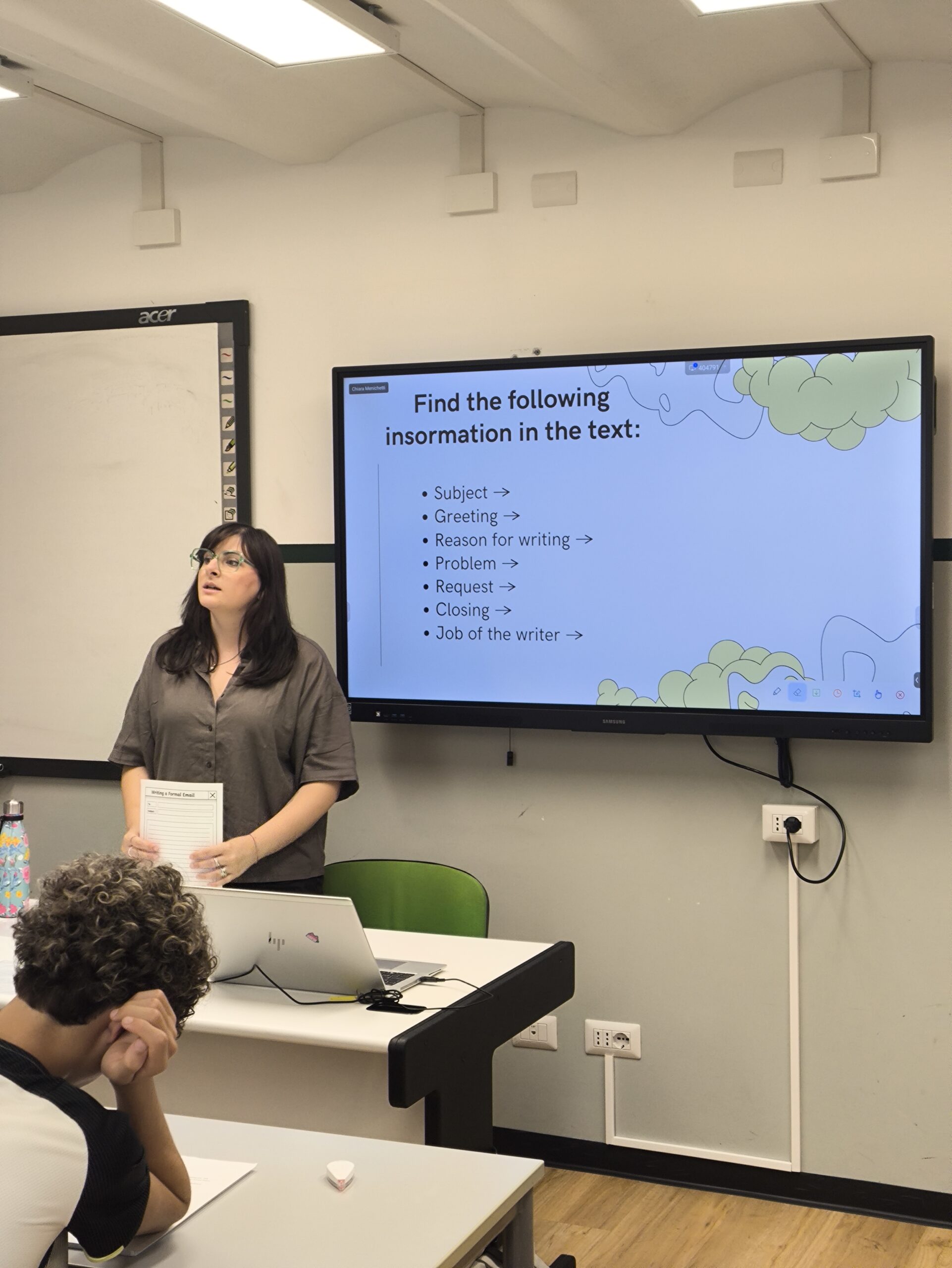 Chiara è in piedi in aula, e dietro di lei ce uno schermo che proietta una slide della lezione di inglese. Ha i capelli di media lunghezza scuri. Porta degli occhiali da vista verdi e un camicia a maniche corte marrone.